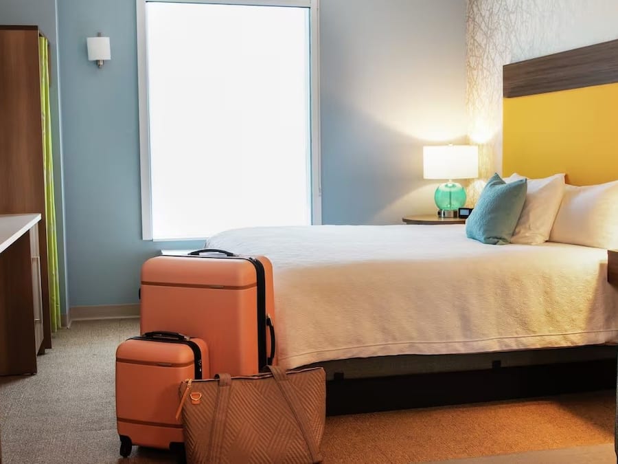 Quarto de hotel com cama, escrivaninha, cadeira, mala e luminária.