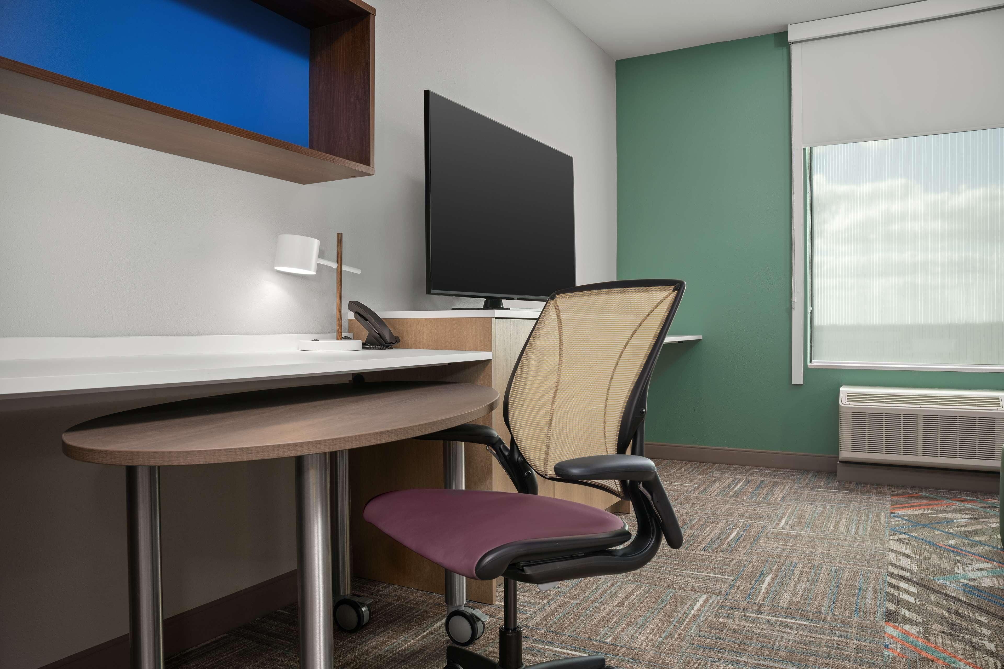 A hotel room with a desk, chair, television, and a window with blinds.