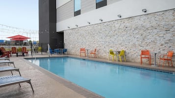 Una piscina con sillas de descanso y mesas, un muro de piedra y un edificio moderno al fondo.