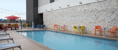 Ein Schwimmbecken mit Liegestühlen und Tischen, eine Steinmauer und ein modernes Gebäude im Hintergrund.