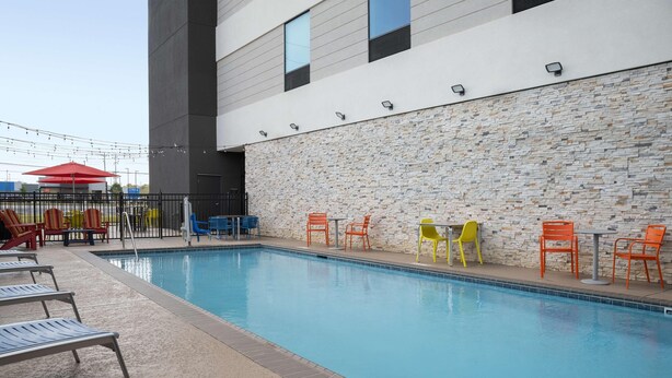 A swimming pool with lounge chairs and tables, a stone wall, and a modern building in the background. - Home2 Suites By Hilton Cullman (Cullman)