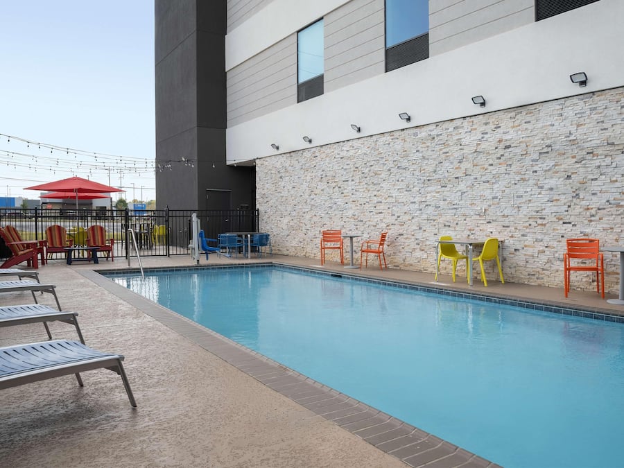 Una piscina con sillas de descanso y mesas, un muro de piedra y un edificio moderno al fondo.