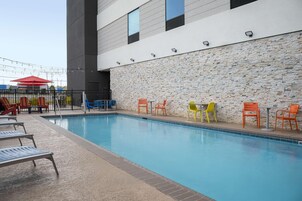Una piscina con sillas de descanso y mesas, un muro de piedra y un edificio moderno al fondo.