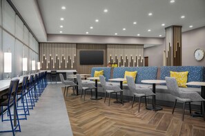 A modern restaurant with long tables and booths, a large TV screen, and a clock on the wall. - Home2 Suites By Hilton Cullman (Cullman)