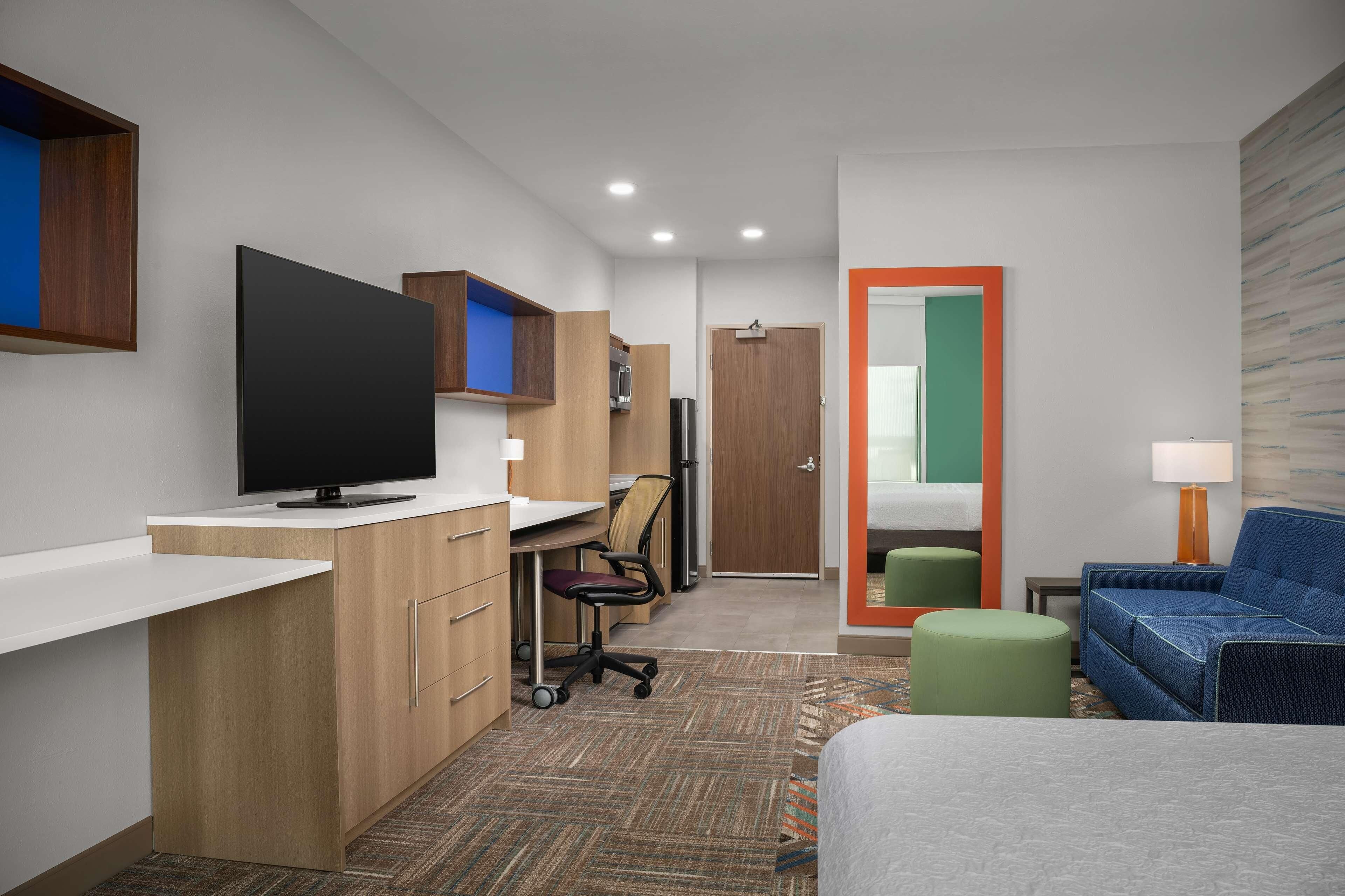 A modern hotel room with a flat-screen TV, a desk with a chair, a sofa, and a small table with a lamp.