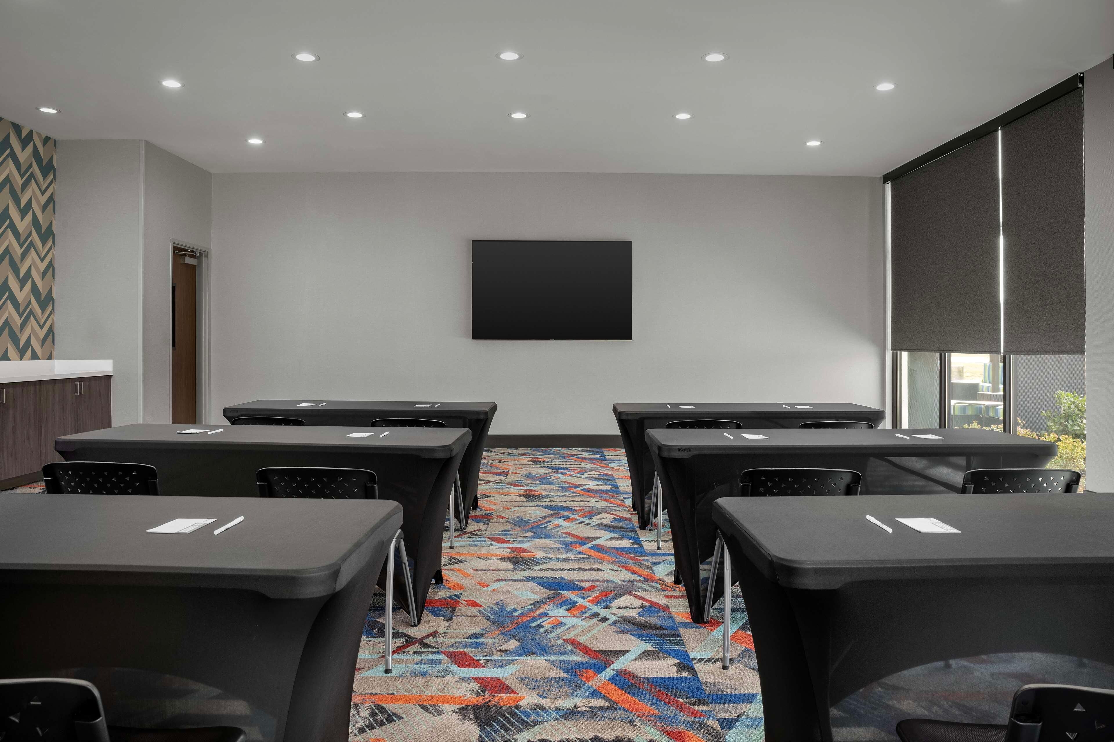 Sala de conferência com tela grande, várias mesas, cadeiras, piso coberto por carpete e uma janela com persianas.