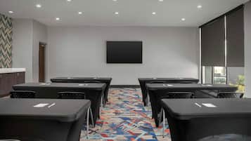 Sala de conferência com tela grande, várias mesas, cadeiras, piso coberto por carpete e uma janela com persianas.