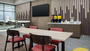 A modern dining area with a long table, red upholstered chairs, a flat-screen TV, and a coffee station with branded dispensers. - Home2 Suites By Hilton Cullman (Cullman)