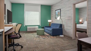 A hotel room with a desk, chair, sofa, ottoman, and a bed. - Home2 Suites By Hilton Cullman (Cullman)