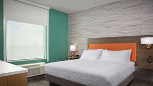 A hotel room with a large bed, a desk, a window with blinds, a wall-mounted lamp, and a heater. - Home2 Suites By Hilton Cullman (Cullman)