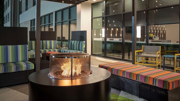 Un área de estar moderna al aire libre con mobiliario de mimbre, una mesa redonda con chimenea y grandes ventanales que ofrecen vistas de la ciudad.