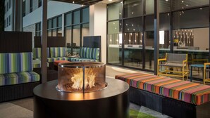 A modern outdoor seating area with wicker furniture, a round table with a fire feature, and large windows offering views of the city. - Home2 Suites By Hilton Cullman (Cullman)
