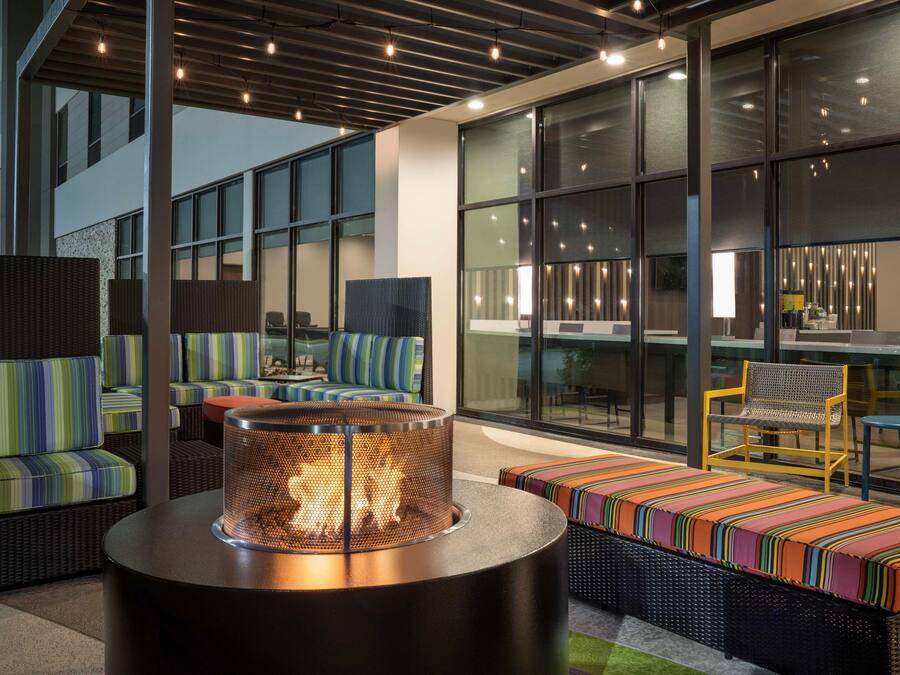 A modern outdoor seating area with wicker furniture, a round table with a fire feature, and large windows offering views of the city.