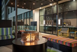 Un área de estar moderna al aire libre con mobiliario de mimbre, una mesa redonda con chimenea y grandes ventanales que ofrecen vistas de la ciudad.