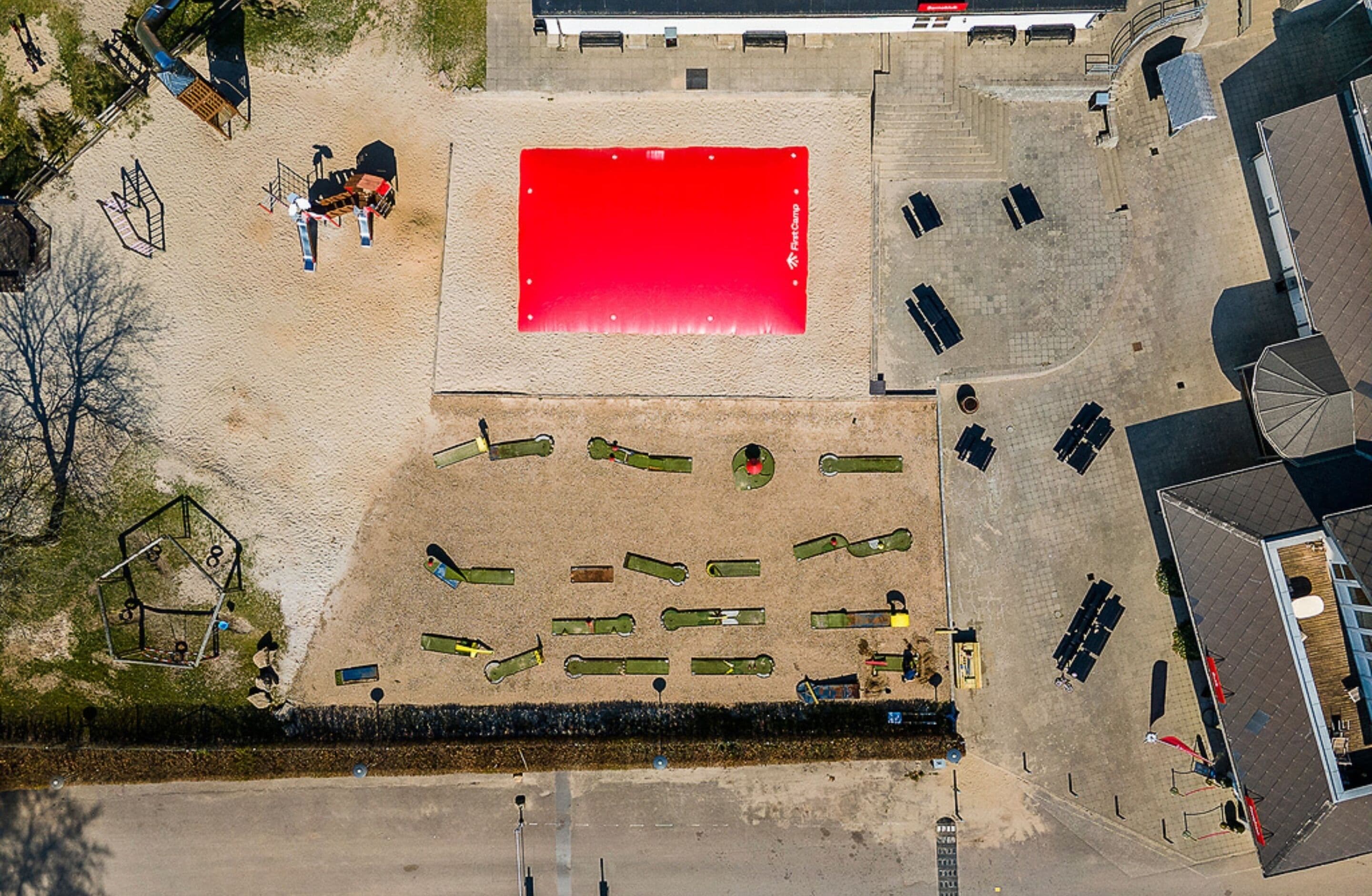 Ein Luftbild eines Parkplatzes mit einem roten, rechteckigen Bereich, möglicherweise ein Sportplatz oder eine Spielwiese, umgeben von geparkten Autos und einigen Personen.