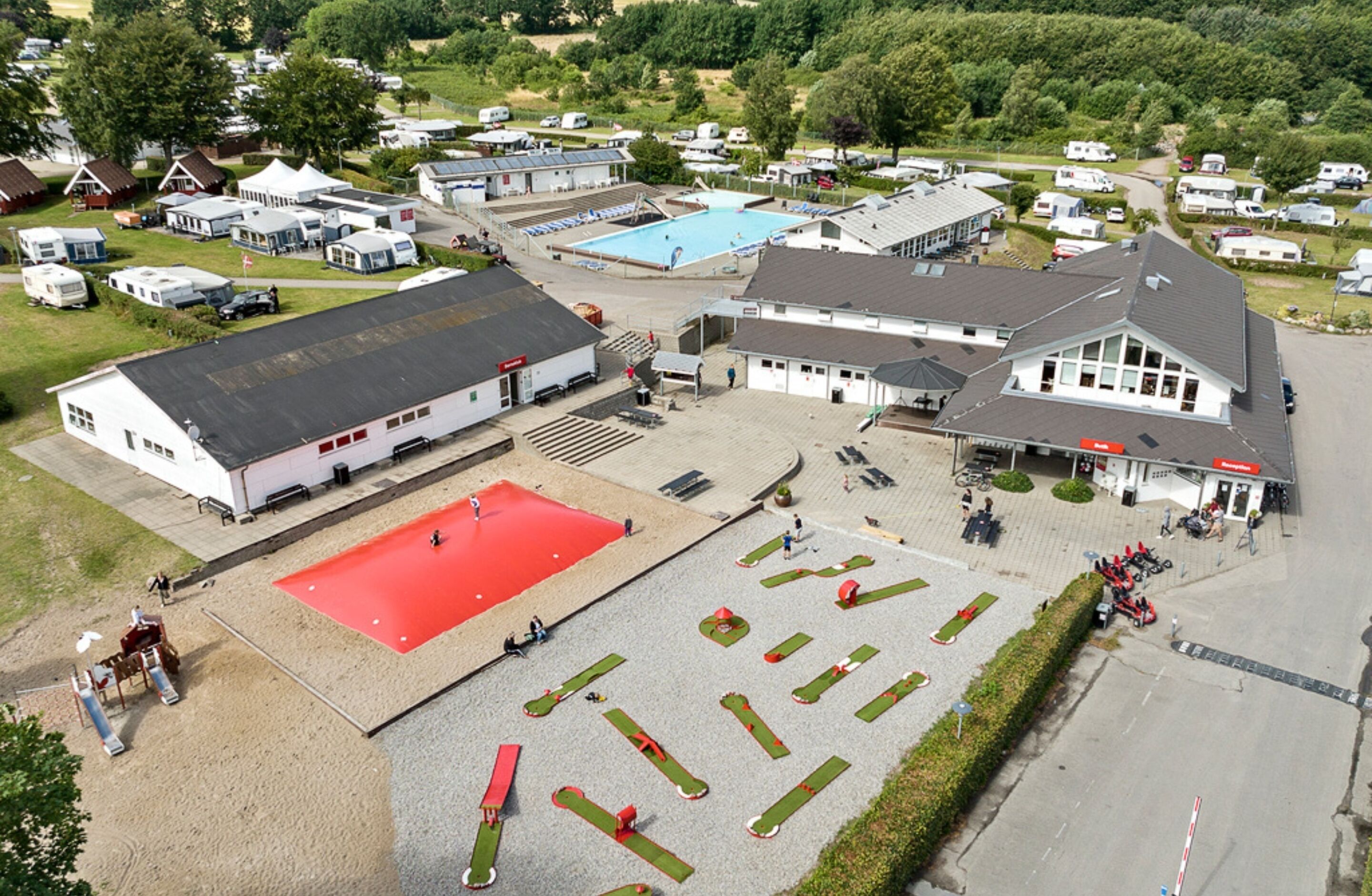 Vista aérea de un camping con vehículos recreativos, una piscina y un edificio grande con techo rojo.