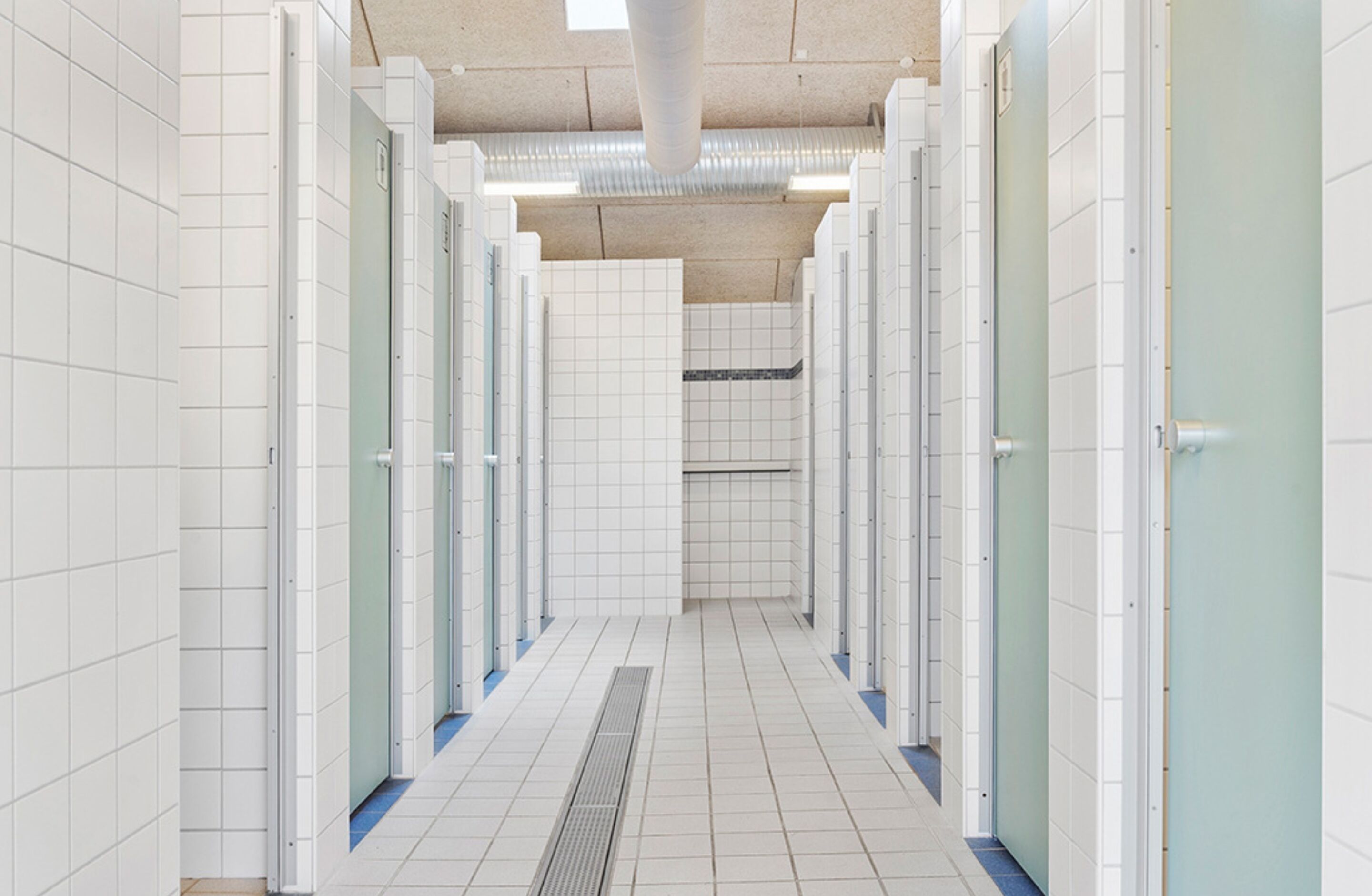 A clean, tiled restroom corridor with multiple stalls, a ventilation system, and a drain in the center.