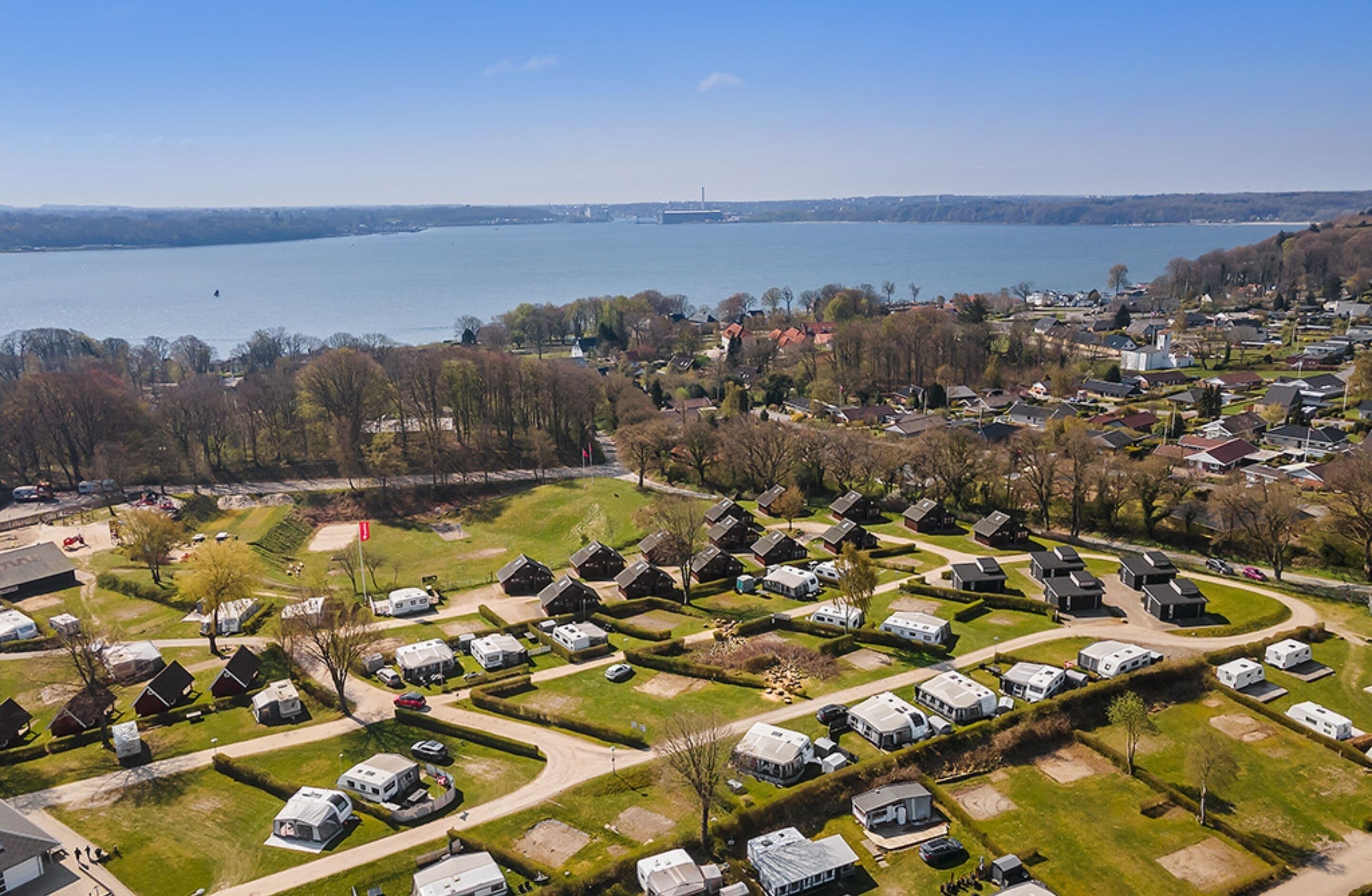 Vista aérea de un camping con vehículos recreativos y casas móviles, rodeado de árboles y un lago.