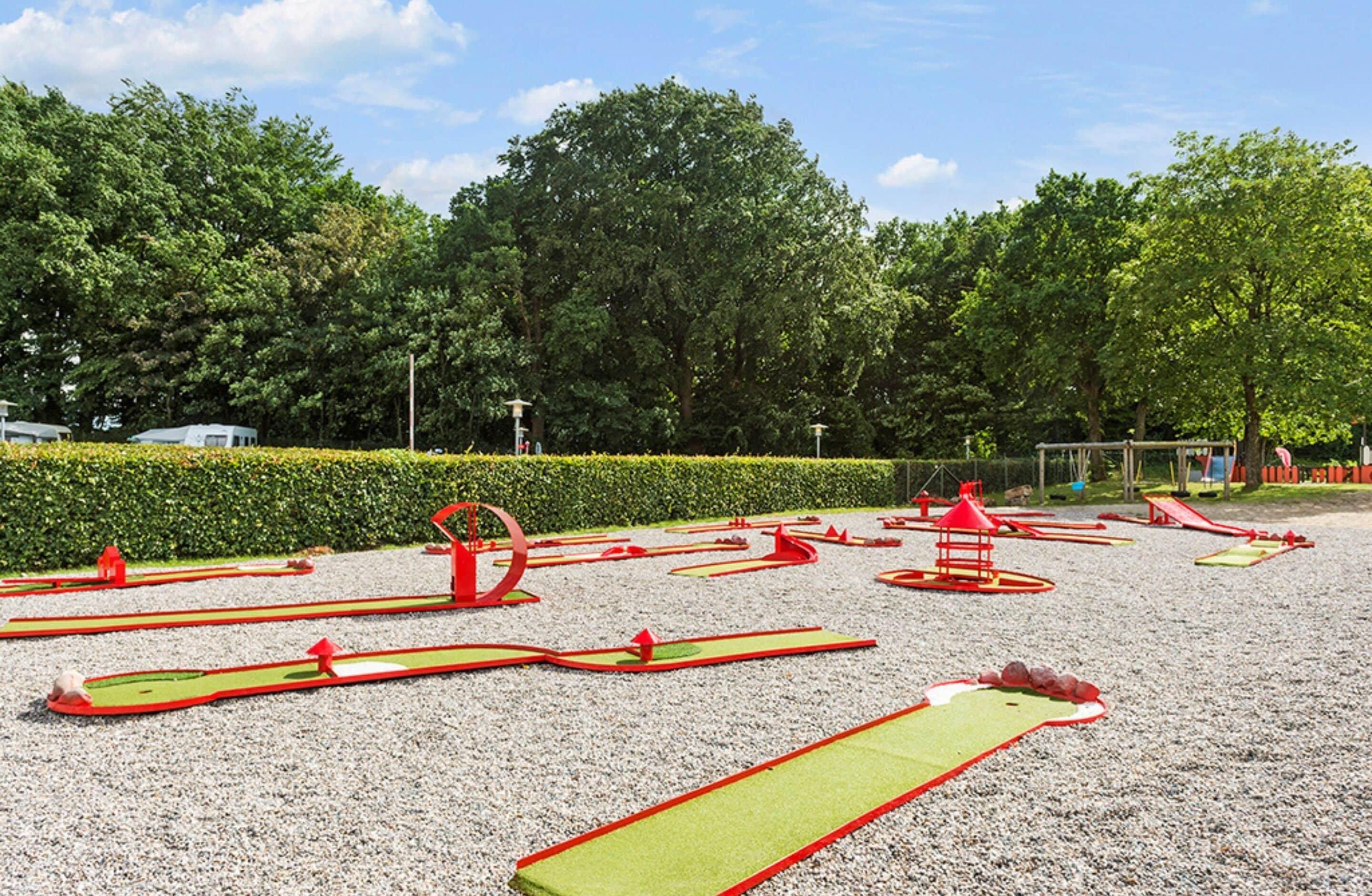 Un espace de jeux extérieur avec différentes structures colorées, principalement en rouge et en vert, installé sur un sol de gravier et entouré d’arbres et d’une haie.