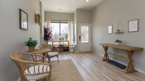Um quarto espaçoso com área de jantar, móveis de vime e uma mesa de madeira, uma janela grande com cortinas, e uma porta que dá para outro cômodo.