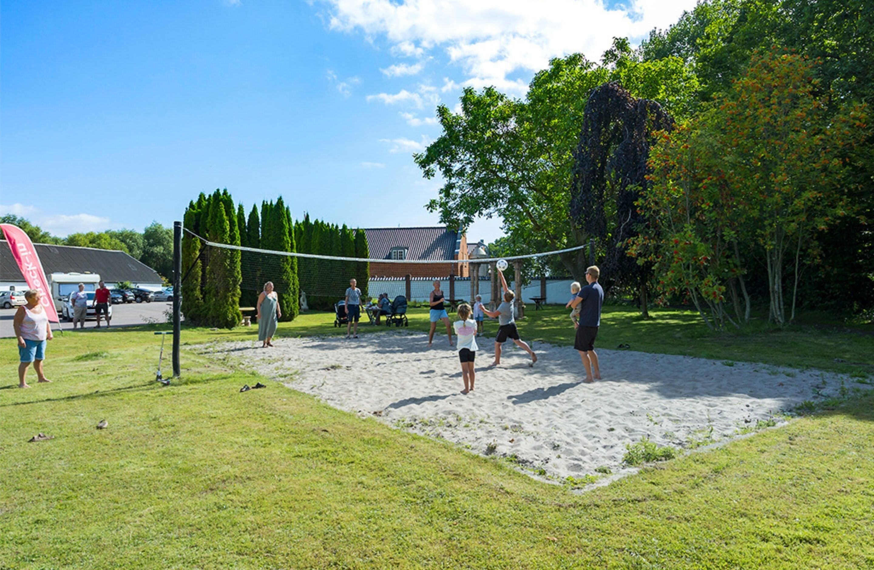 A volleyball game on a sandy court with a grassy area in the foreground, surrounded by trees and a building in the background.