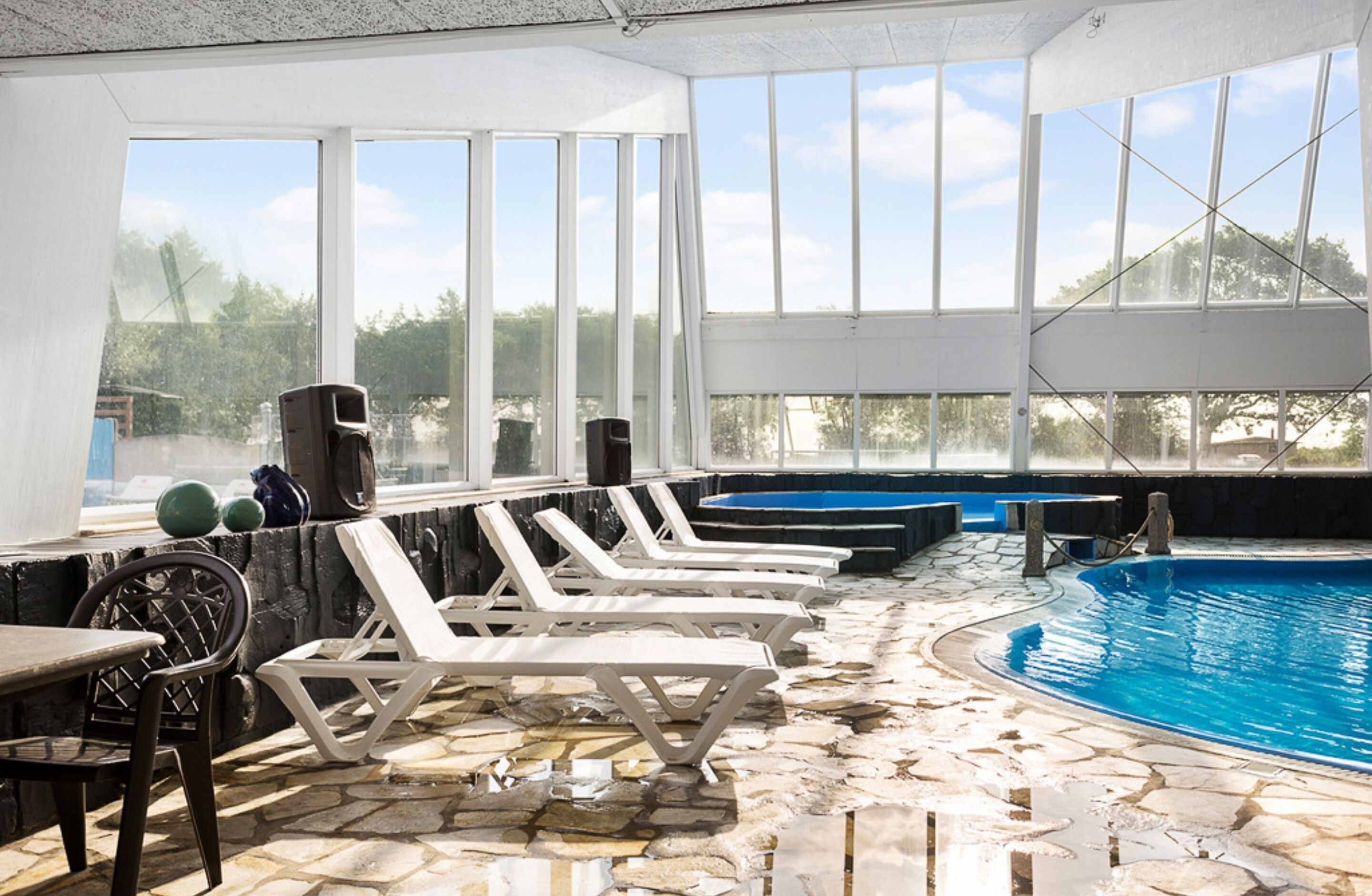 An indoor swimming pool with stone flooring, white lounge chairs, and large windows offering a view of the outdoors.