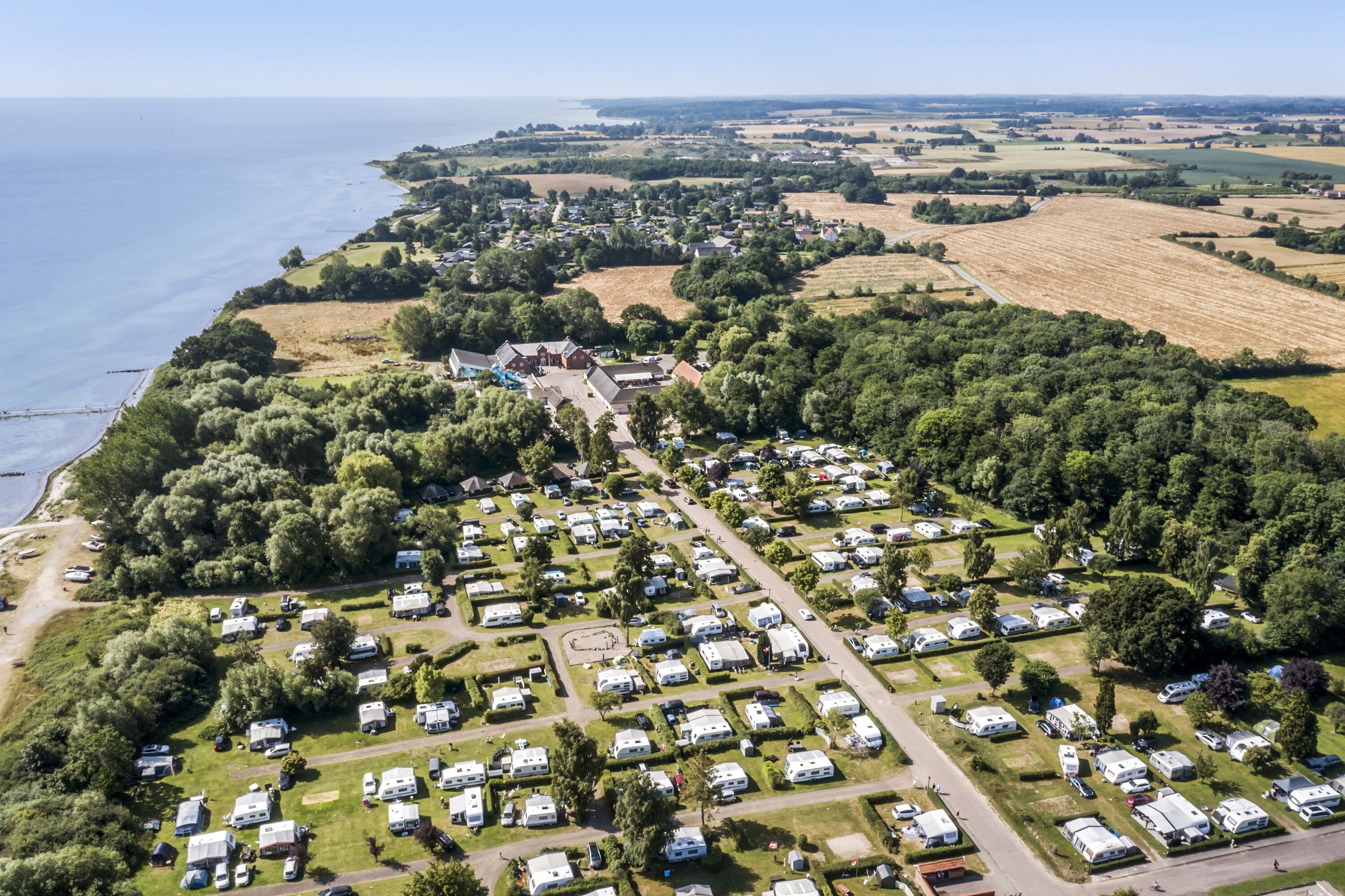 An aerial view of a campsite with RVs and tents arranged in rows, surrounded by greenery and fields, with a body of water in the background.