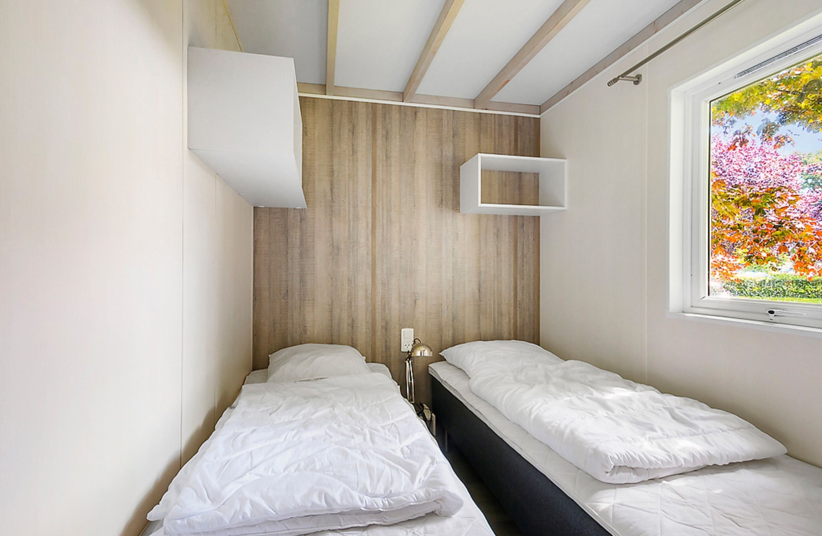 Un dormitorio con dos camas, un cabecero de madera, una ventana con vistas a árboles y un estante blanco.