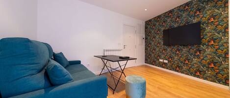 A modern living room with a blue sofa, a small table, a round ottoman, and a wall-mounted TV.