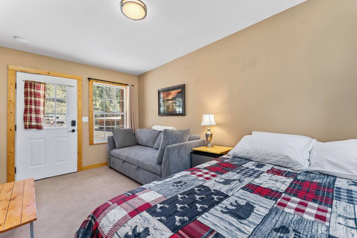 A bedroom with a bed, a grey sofa, a wooden table, a lamp, and a door with curtains.