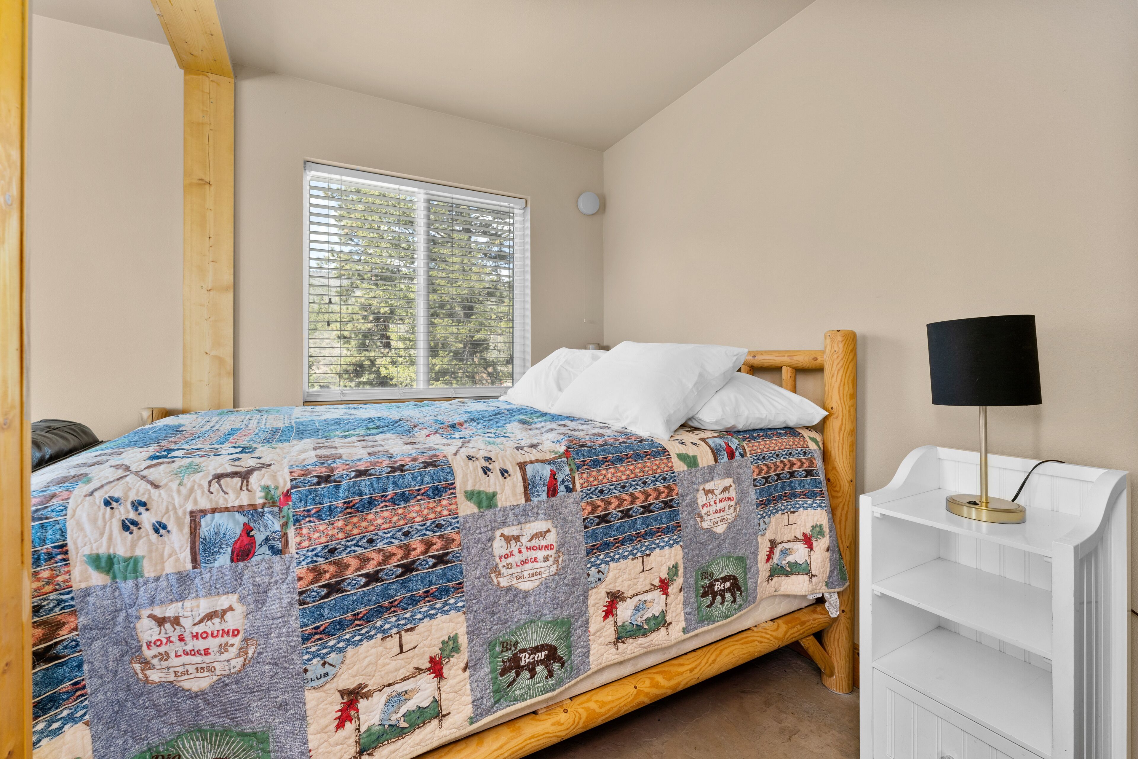 A bedroom with a wooden bed, a quilt with animal prints, a window with blinds, and a white nightstand with a lamp.