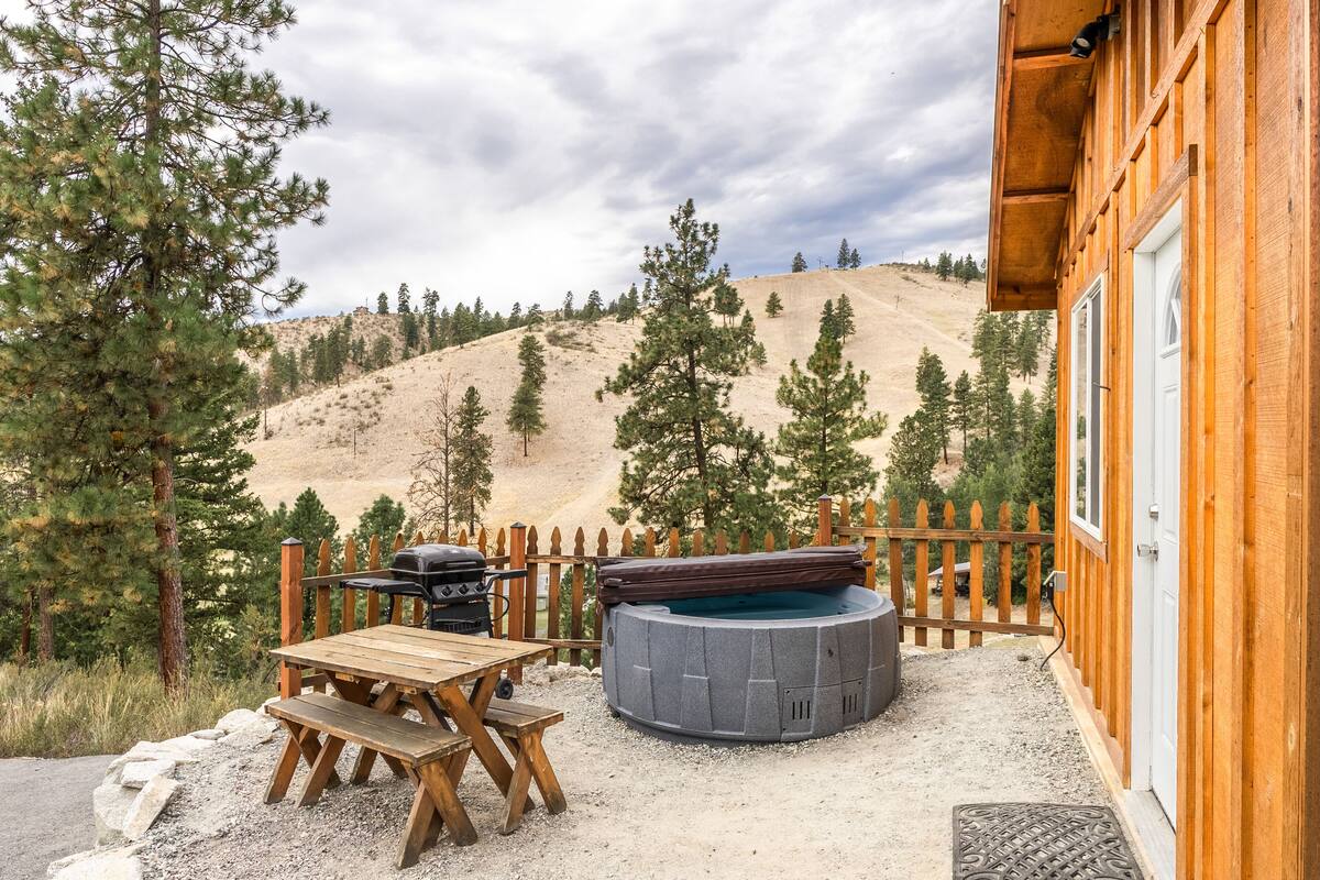 A wooden cabin with a hot tub, a barbecue grill, and a picnic table in a mountainous area.