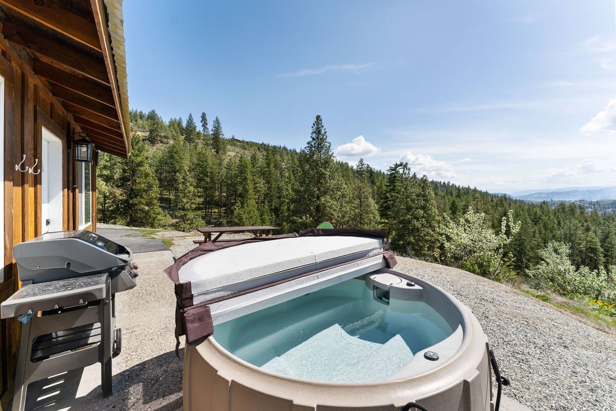 A hot tub with a wooden deck, a barbecue grill, and a view of a forested hillside.