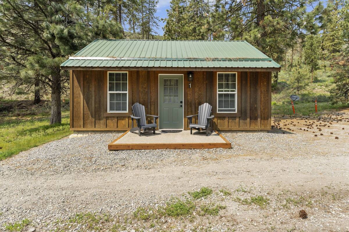 A wooden cabin with a green roof, two Adirondack chairs outside, and a gravel driveway.