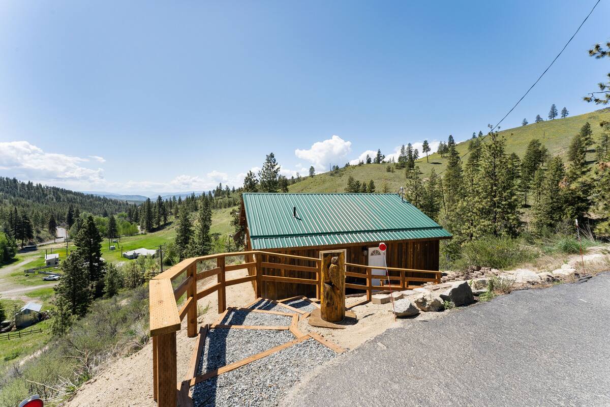 A wooden cabin with a green roof, surrounded by a wooden fence, set against a backdrop of a mountainous landscape.
