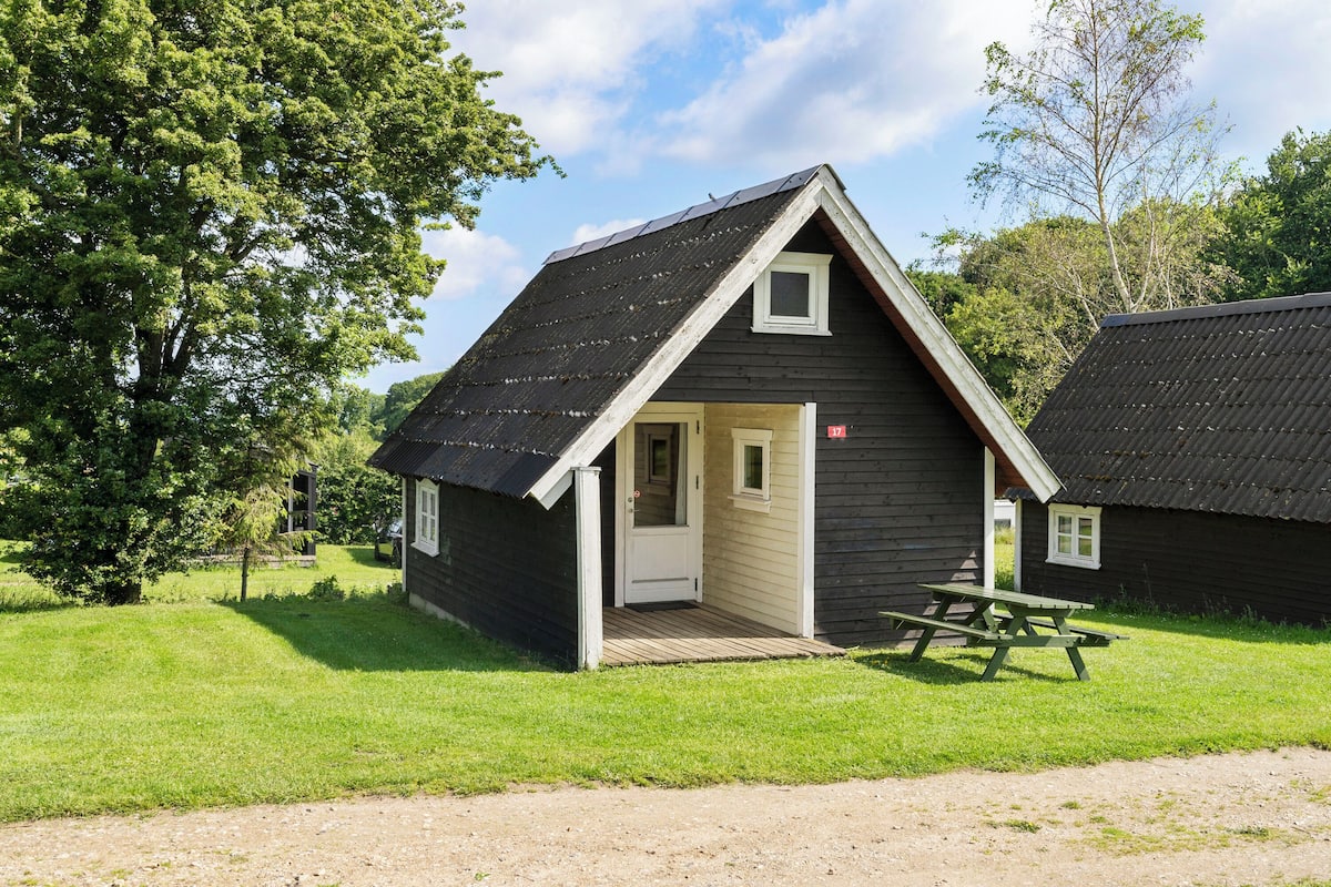 A small, dark wooden cabin with a sloped roof, a door, and a window. There is a picnic table in front of the cabin.