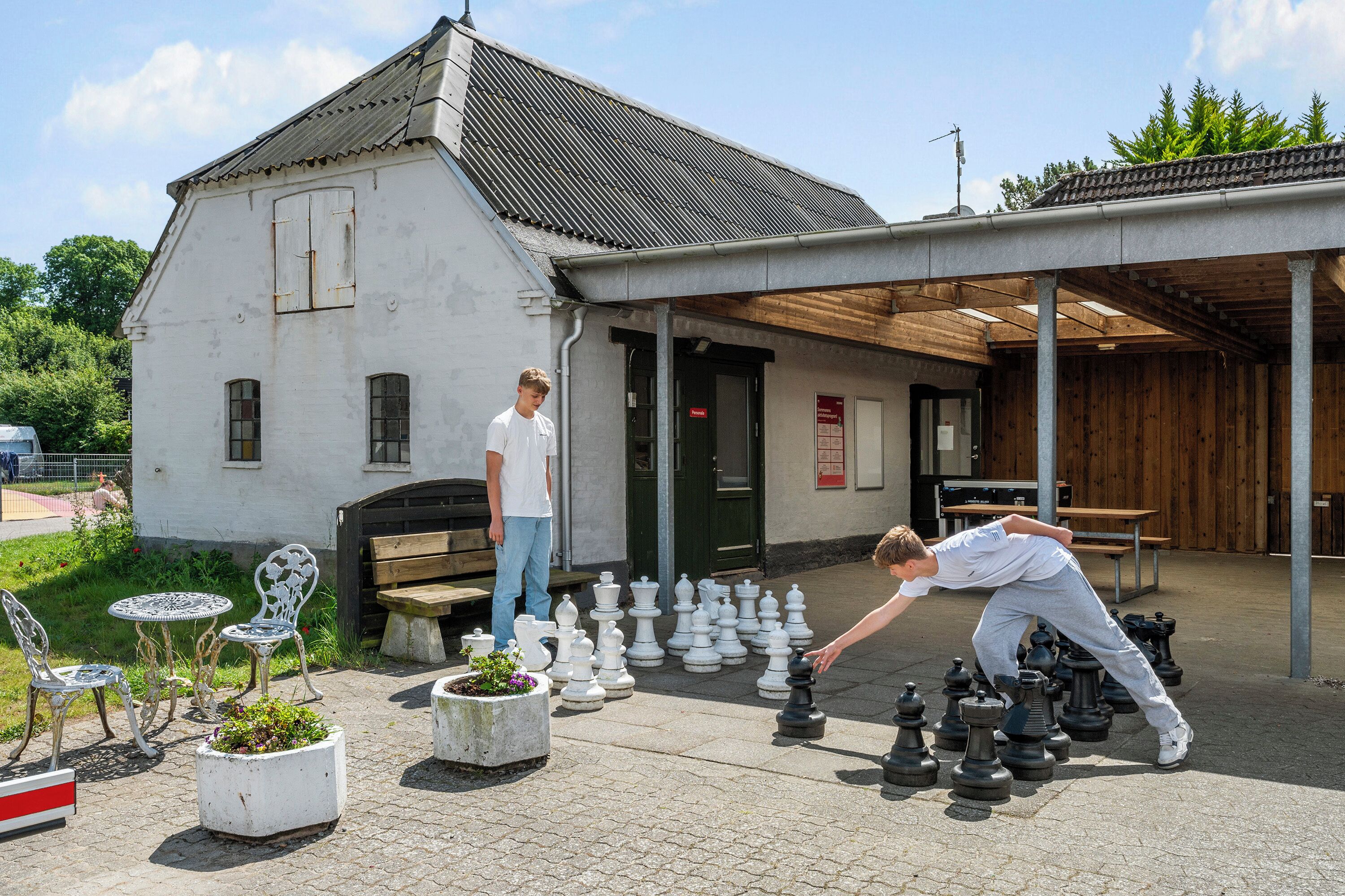 An outdoor chess setup with multiple chess pieces arranged on a paved surface, a white building with a sloped roof in the background, and a wooden bench.