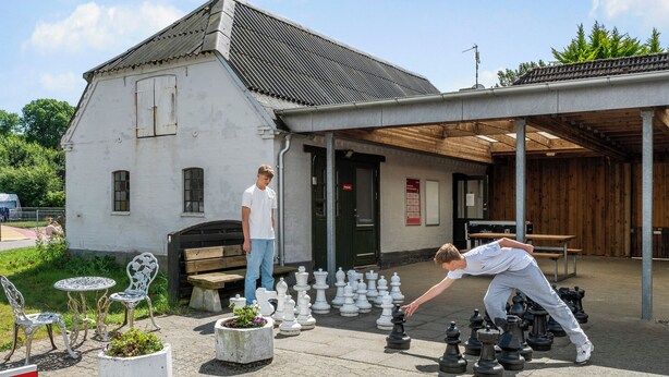 An outdoor chess setup with multiple chess pieces arranged on a paved surface, a white building with a sloped roof in the background, and a wooden bench. - First Camp Skovlund  Lillebælt (Middelfart)
