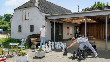 An outdoor chess setup with multiple chess pieces arranged on a paved surface, a white building with a sloped roof in the background, and a wooden bench.