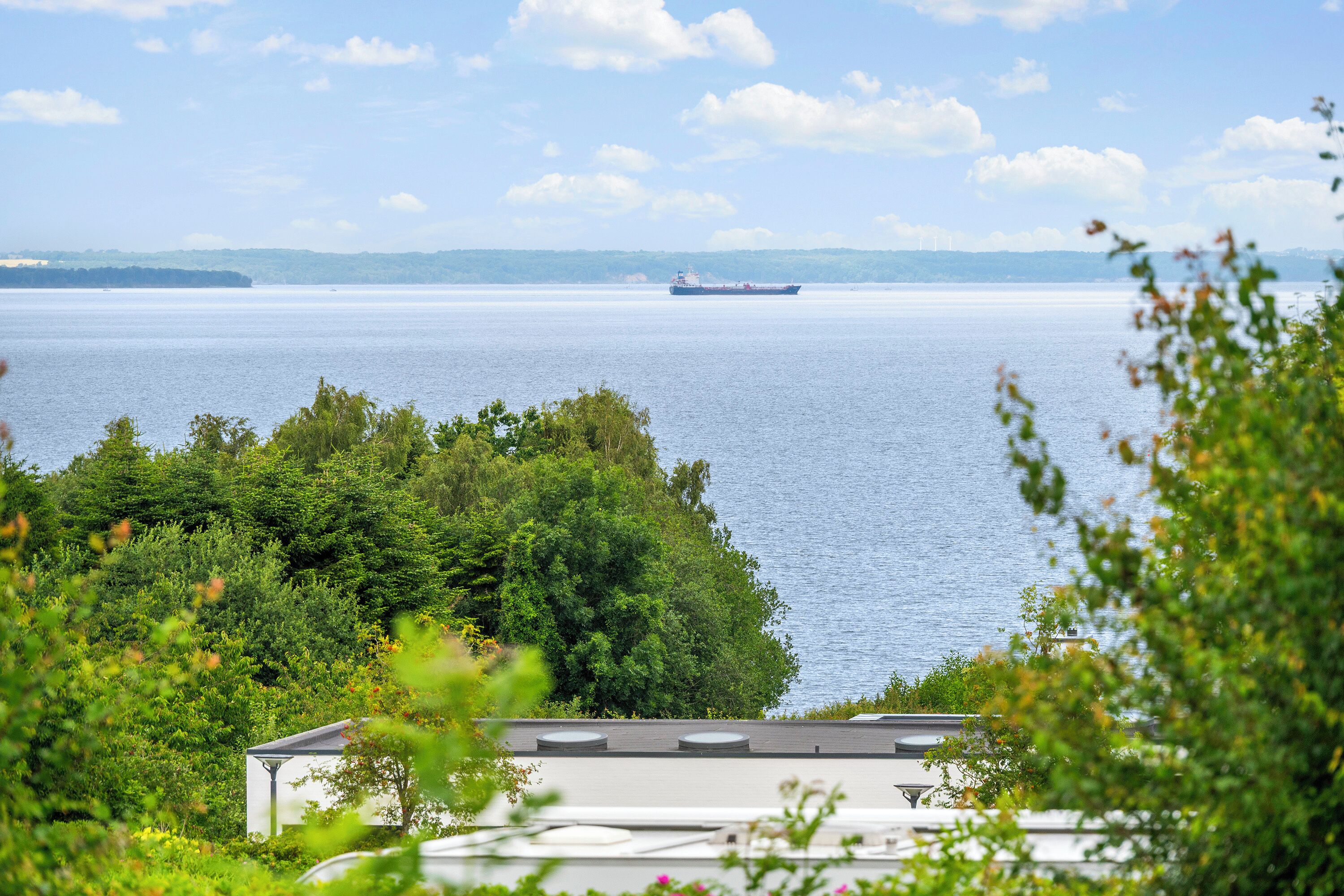 A large body of water with a ship in the distance, surrounded by dense green foliage and a building with a flat roof.