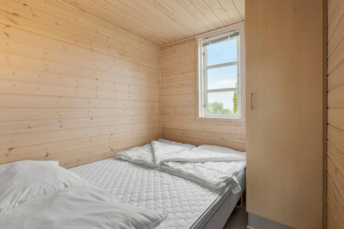 A small, wooden-paneled room with a single bed, a window, and a door.