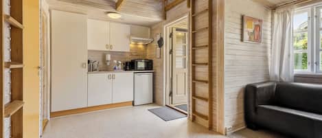 Una cocina compacta con armarios blancos, un refrigerador negro y un microondas. Una escalera de madera está apoyada contra la pared y hay un sofá de cuero negro en el área de estar contigua.