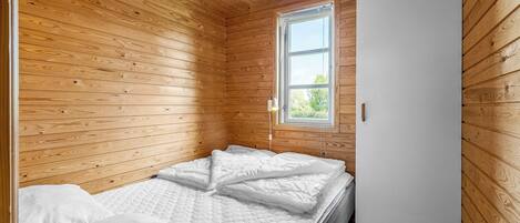 A small, wooden-paneled room with a single bed, a window, and a door.