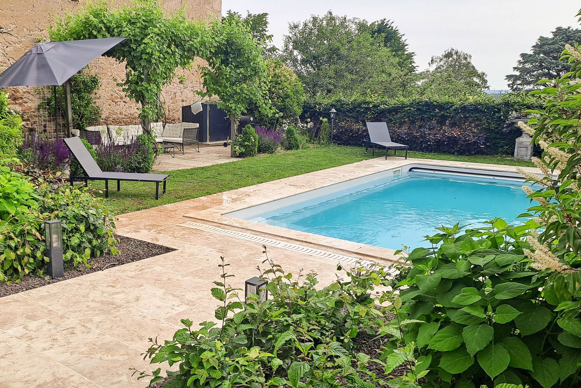 Una piscina rectangular con solana empavedada, rodeada de abundante vegetación y un muro de piedra.
