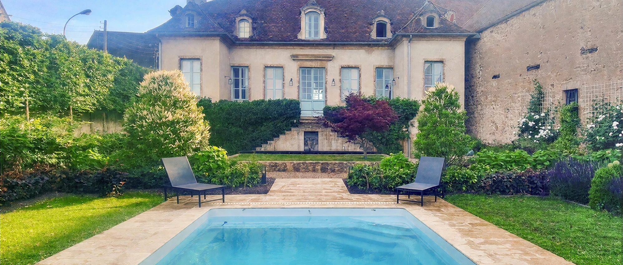 A swimming pool with two lounge chairs, surrounded by a well-maintained garden and a large building with multiple windows.