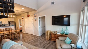 A spacious living area with a wooden floor, a dining table with chairs, a sofa, a television mounted on the wall, and a window providing natural light.
