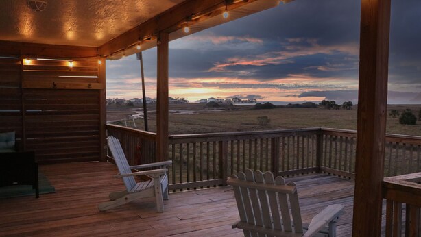 A wooden deck with two Adirondack chairs and a view of a sunset over a grassy field.