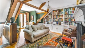 A cozy living room with a high ceiling, exposed wooden beams, a fireplace, a bookshelf filled with books, and a comfortable sofa.