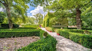 A garden pathway leading to a stone building surrounded by neatly trimmed hedges and trees.