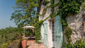 A rustic outdoor patio with a table and chairs, a large potted plant, and a partially open door to a building with ivy-covered walls.